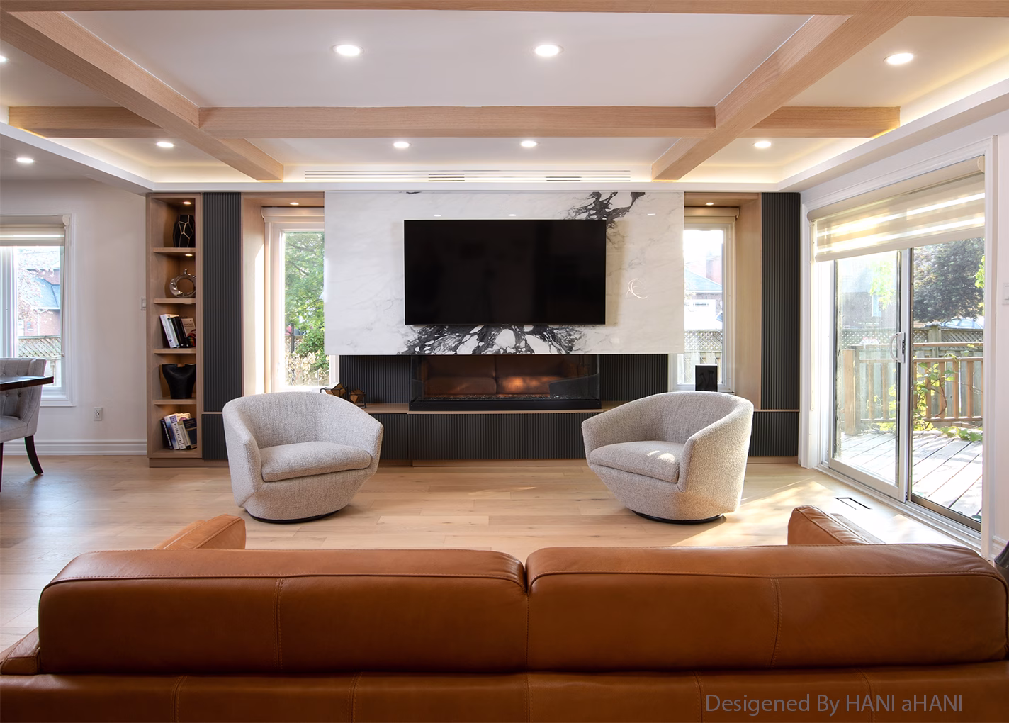 A sophisticated living room features a modern fireplace area with a large flat-screen TV mounted above it, set against a stunning white marble slab with prominent dark veining. Below the fireplace, dark fluted panels run horizontally. On either side of the fireplace, two light grey swivel chairs face the central feature. To the left, a dark-toned built-in shelving unit with open shelves displays books and decorative items, flanked by a window. The ceiling has a contemporary design with light wood beams and recessed lighting. The room is floored with light-colored wood. In the foreground, the back of a brown leather sofa is visible. On the right, large sliding glass doors lead to an outdoor space, allowing natural light to fill the room.